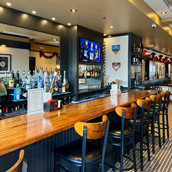 A long, polished wooden bar with several stools, liquor bottles, and a TV showing a sports game.