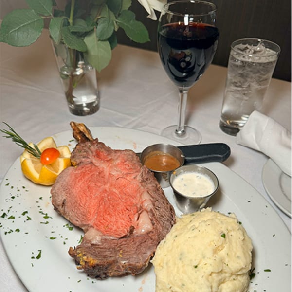 Prime rib with mashed potatoes, sauces, and a lemon garnish, alongside wine and water glasses.
