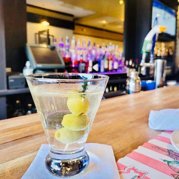 Martini with three olives on a bar counter with blurred bottles in the background at Spaghettini Pizza Trattoria.