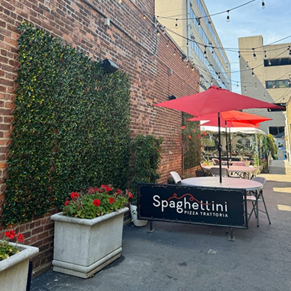 Outdoor patio of Spaghettini Pizza Trattoria with red umbrellas, tables, brick wall, and string lights.