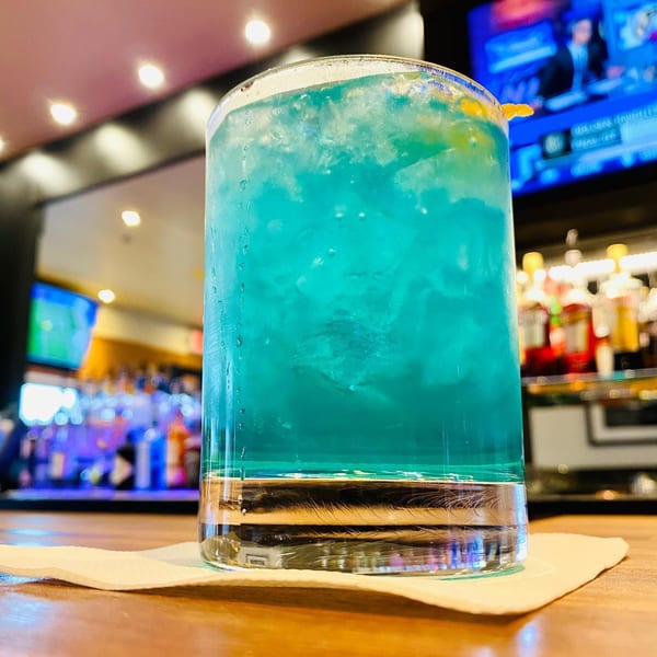 A vibrant blue cocktail with ice and an orange garnish sits on a bar with blurred bottles in the background.