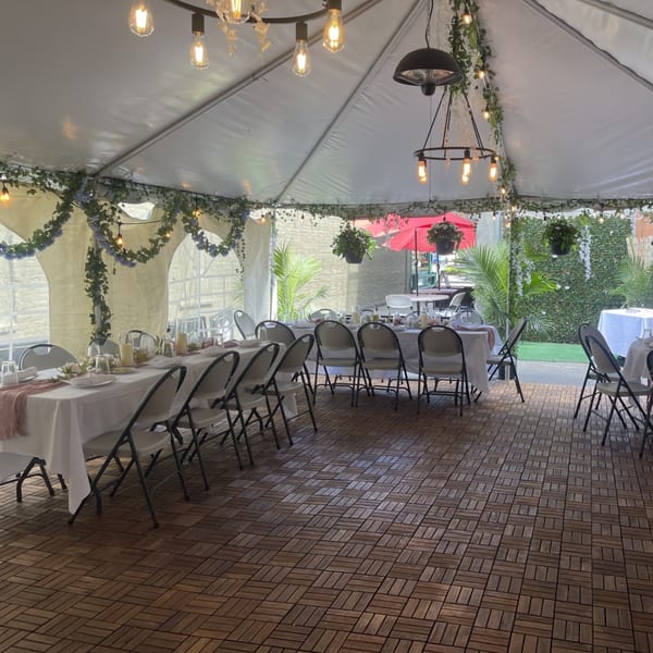 Outdoor dining area under a white tent with string lights, set tables, and green foliage.