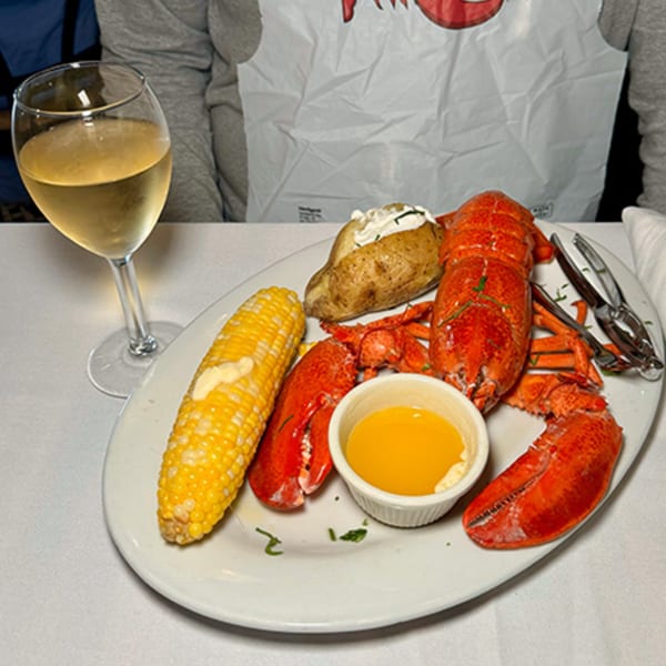 A plate of cooked lobster, corn on the cob, baked potato, and melted butter with a glass of white wine.