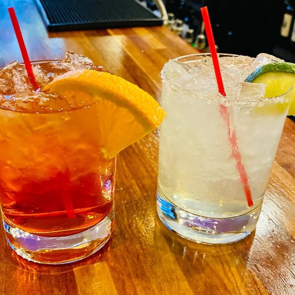 Two cocktails, one orange with an orange slice, one clear with lime, on a wooden bar.